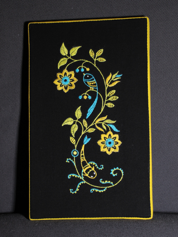 Embroidery in neon yellow, blue and green on black fabric, showing a bird and a fish as part of a floral swirl pattern. It is mounted apparently-seamlessly on a stretcher, with a tidy yellow border sewn over the edge.