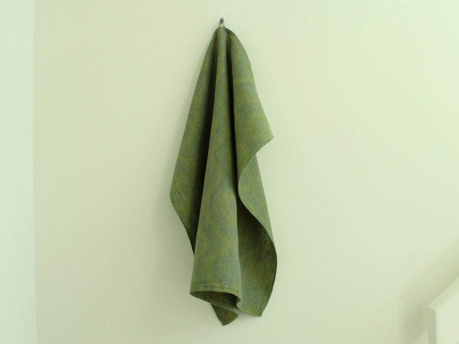 A green towel with a subtle ring pattern hanging from a nail on a white wall, showcasing that it drapes neatly.