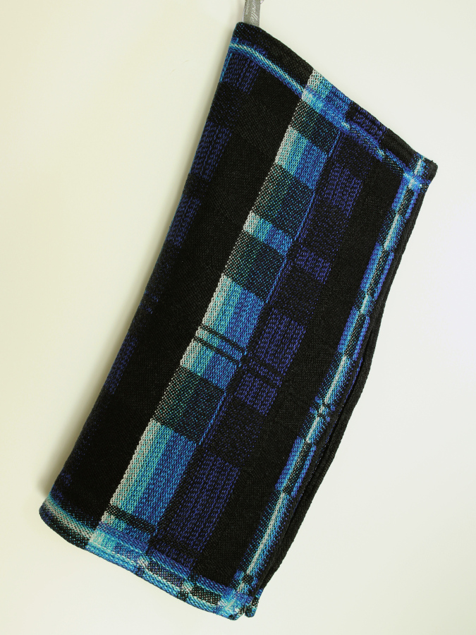 A black towel with neon blue borders and patterns, forming irregular checks, hanging from an unseen nail and folded in half as a result.