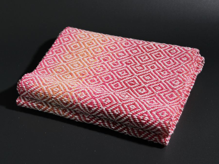 A scarf in pink-and-orange wool, with irregular diamond patterns in white, neatly folded.