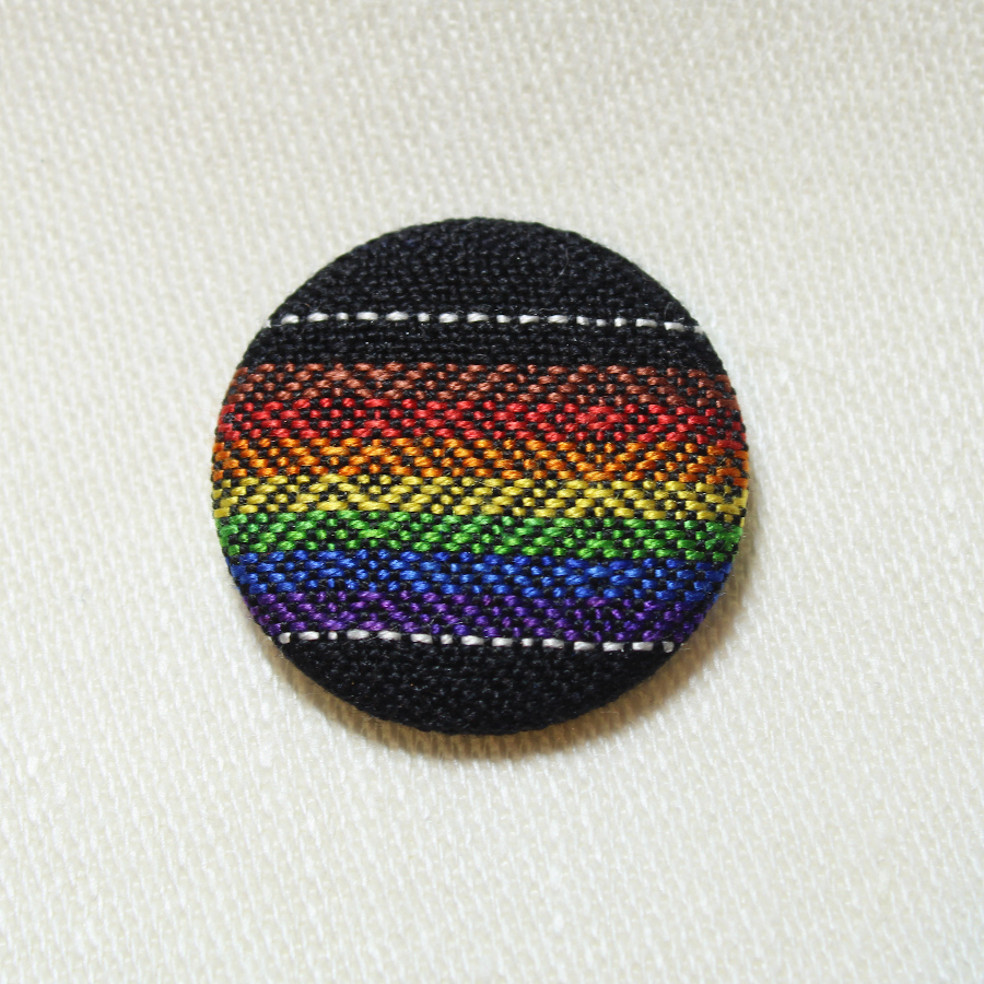 A fabric-covered pinback button, showing the Philadelphia Pride flag in shiny silk with subtle diamond patterns on black.