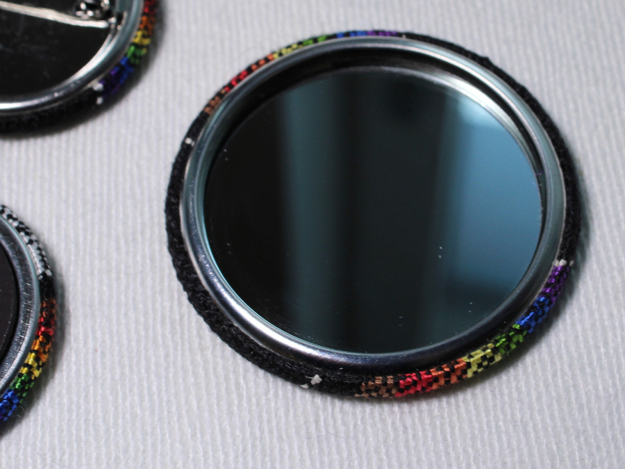 A fabric-covered pocket mirror, turned upside-down to show the mirror side. The edges show that the fabric is the Philadelphia Pride flag on black.