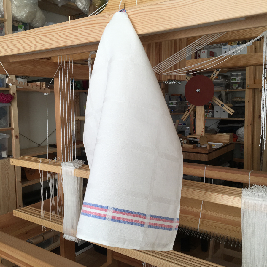 A white linen towel with a trans Pride flag stripe, hanging from a nail at the front of a wooden loom.