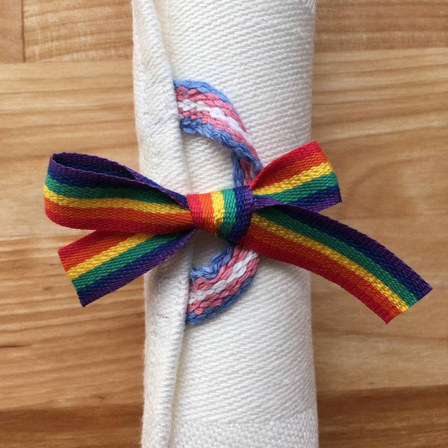 A white linen towel with a trans Pride flag hang loop, rolled up neatly and held so by a bow of rainbow ribbon.