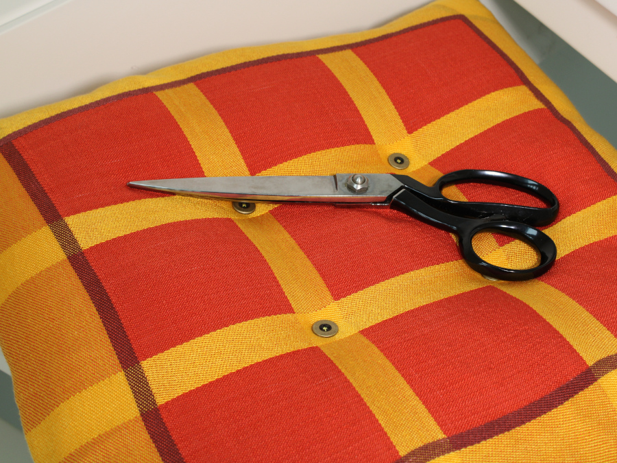 A square cushion with a thick yellow border and a three-by-three grid pattern of red squares, cinched by buttons and looking quite squishy. A pair of sewing shears lies in the middle.